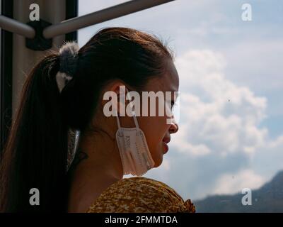 Medellin, Antioquia, Kolumbien - März 27 2021: Junge Asiatin mit einer Gesichtsmaske unter ihrem Mund schaut durch das Fenster Stockfoto