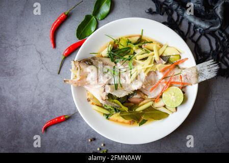 Gedünsteter Zackenbarsch mit Sojasauce nach chinesischer Art, gedünsteter Fisch auf weißem Teller mit Ingwer-Chili-Kraut und Gewürzen Zitronenlime auf dunklem Hintergrund Stockfoto