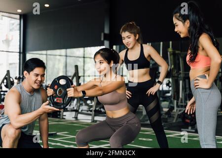 Gruppe von Menschen jubeln auf ihre asiatische Freundin tun Kniebeugen mit einer Hantelscheibe in Fitness-Studio. Gemeinsam als Teamwork arbeiten. Ermutigung und Zusammengehörigkeitskonzept Stockfoto