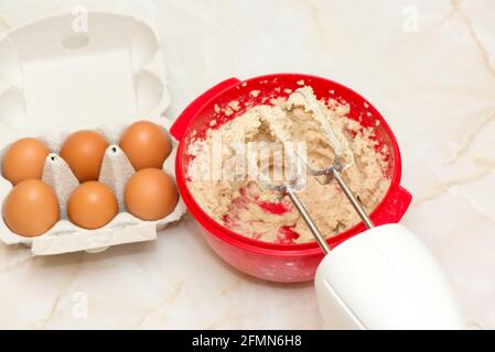 Elektrischer Quirl auf einer roten Schüssel mit Teig und Ei Karton Stockfoto