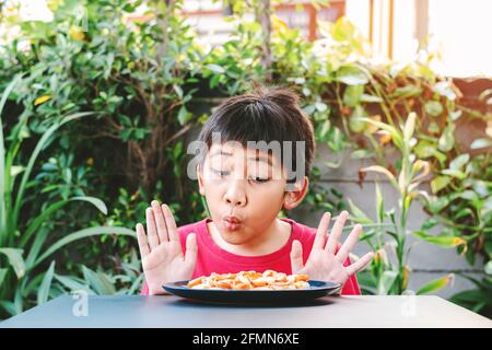 Ein süßes asiatisches Kind in einem roten Hemd zeigte einen guten Ausdruck, als er eine Pizza auf einem Teller vor dem Tisch sah. Stockfoto