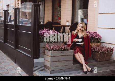 Schöne junge blonde Frau in einem trendigen Herbst-Outfit in burgunderrot-marsala Farbe mit einer Tasse Kaffee auf den Stufen eines Cafés. Weicher, selektiver Fokus. Ar Stockfoto