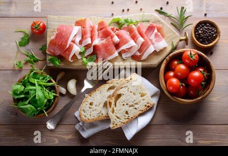 Rustikales Gericht, roher Schinken auf Schneidebrett mit Brot, Rucola und Kirschtomaten. Draufsicht auf Holzhintergrund. Stockfoto