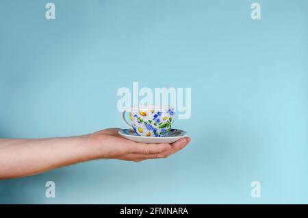 Hand und Tasse auf blauem Hintergrund. Mann hält weiße Tasse mit bemalten Blumen. Tasse aus Keramik und Untertasse für Tee oder Kaffee hell in Blau A dekoriert Stockfoto