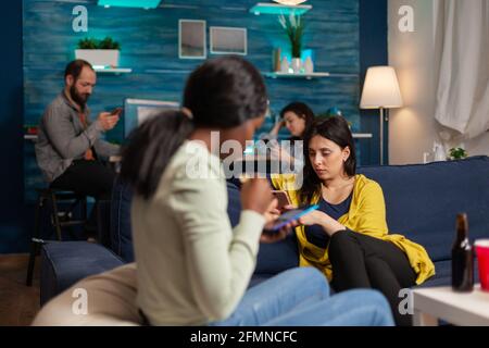 Mixed-Rennen Freunde sitzen auf Sofa-Bestände mobile in Händen Surfen im Internet, Online-Videos während einer entspannten Party zu sehen. Eine Gruppe multiethnischer Menschen, die gemeinsam im Wohnzimmer herumhängen Stockfoto