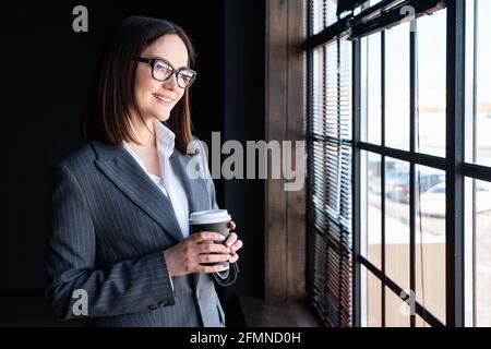 Eine junge Frau in einem Geschäftsanzug mit einem Getränk in einem Papierglas blickt aus dem Fenster. Stockfoto