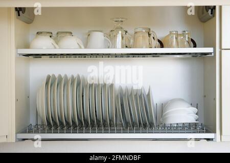 Regal zum Trocknen von Geschirr in der modernen Küche. Weiße Glas- und Keramikplatten und -Tassen auf Metallregal im Küchenschrank. Stockfoto