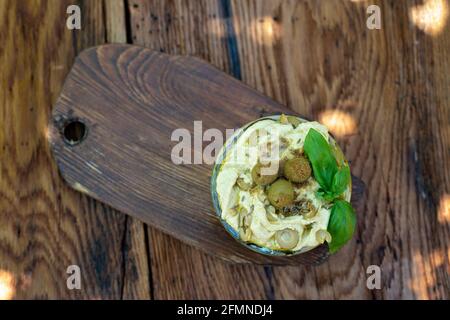 Klassischer Appetizer-Hummus mit Oliven und einem Basilikumzweig aus Kichererbsen, Tahini, Olivenöl und orientalischen Gewürzen in einer Schüssel auf einem Holztisch. Herkömmliches M Stockfoto