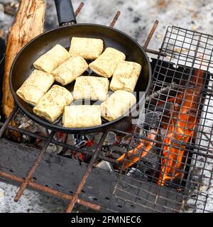 Gerollte Pfannkuchen mit Füllung werden in einer Pfanne auf dem Feuer im Grill gebraten. Winter Kochen Lebensmittel in der Natur im Freien. Traditionelle nationale russische f Stockfoto