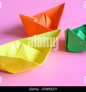 Drei helle bunte Papier Origami Boote in orange, gelb und grün auf einem rosa Hintergrund. Quadrat Stockfoto