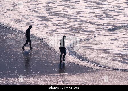 Am Fistral Beach in Newquay in Cornwall laufen zwei Männer im Abendlicht zum Meer, während kleine Wellen sanft fließen. Stockfoto