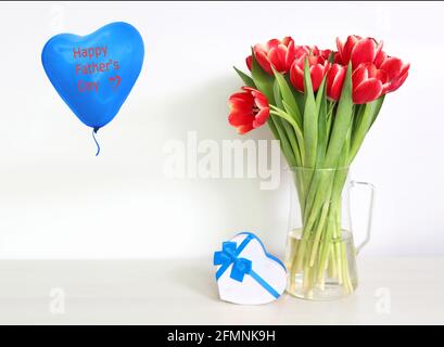 Grußkarte für Herren. Schöne rote Tulpen und eine Geschenkbox in Form eines Herzens auf dem Tisch und einem blauen Ballonherz mit Aufschrift Happy Father‘s Day Stockfoto