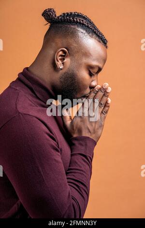 afroamerikanischer Mann mit geschlossenen Augen und zusammen mit den Händen posiert er im Studio vor braunem Hintergrund. Stockfoto