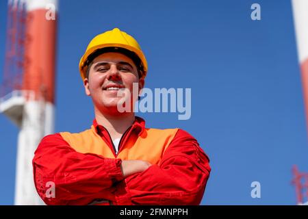 Porträt eines jungen Arbeiters in Red Coveralls und Yellow HardHat gegen die Schornsteine des Kraftwerks und den blauen Himmel. Lächelnder Arbeiter in schützender Arbeitskleidung. Stockfoto