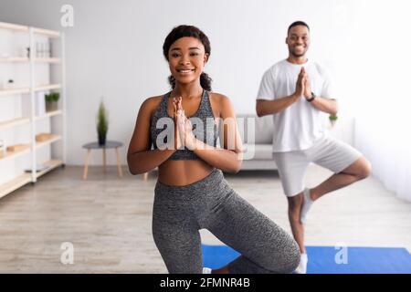 Fröhliche schwarze Frau, die mit ihrem Freund Yoga praktiziert, in Baumhaltung stehend und in Innenräumen eine Namaste-Geste macht Stockfoto