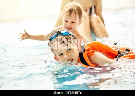 Junge mit Schwanenhals und Sicherheitsweste lernt, in der Nähe zu schwimmen Bruder und Mutter im großen Pool mit klarem Wasser Sportkomplex in der Nähe
