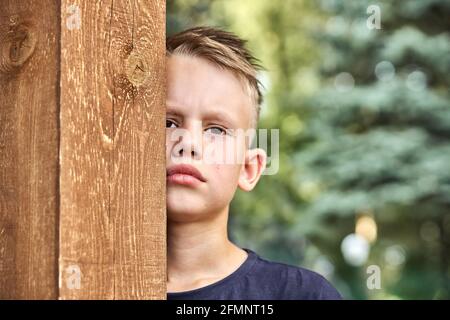 Verärgert blonde Junge trägt dunkle T-Shirt versteckt sich hinter Holzzaun Posiert für die Kamera im grünen sonnigen Sommer Stadtpark in der Nähe Anzeigen Stockfoto