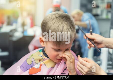 Blonde kleine Junge mit bunten Abdeckung entfernt Haare von der Nase Nach dem Haarschnitt mit einem erfahrenen Stylisten im modernen Schönheitssalon Nahaufnahme Stockfoto