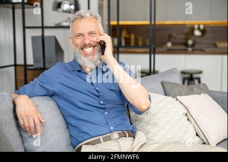 Porträt eines ruhigen älteren bärtigen Kerls, der sich auf der bequemen Couch niedersetzt und auf dem Smartphone spricht, eines reifen Hipsters, der mit Freunden spricht, genießt das Telefongespräch zu Hause und lächelt Stockfoto