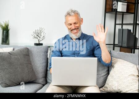 Moderner bärtiger Mann mittleren Alters, der einen Laptop für virtuelle Meetings nutzt, die zu Hause auf der Couch sitzen. Reifer grauhaariger Mann winkt in die Webcam, chattet online, hält Videoanruf Stockfoto
