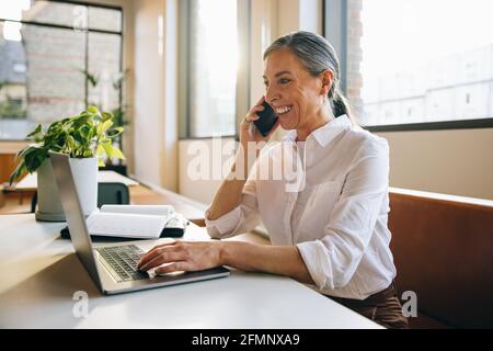 Eine reife Geschäftsfrau am Schreibtisch, die einen Laptop benutzt und mit einem Mobiltelefon spricht. Freiberuflicher Mitarbeiter, der mit dem Kunden über ein Smartphone spricht, während er einen Laptop verwendet. Stockfoto