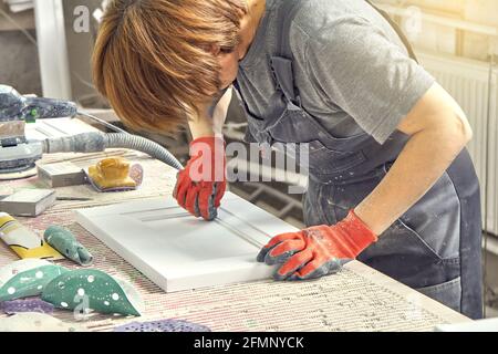 Weibliche Angestellte in schmutziger Uniform und Arbeitshandschuhen poliert Holz Ein Teil der Möbelfassade an der Werkbank in der Werkstatt Stockfoto
