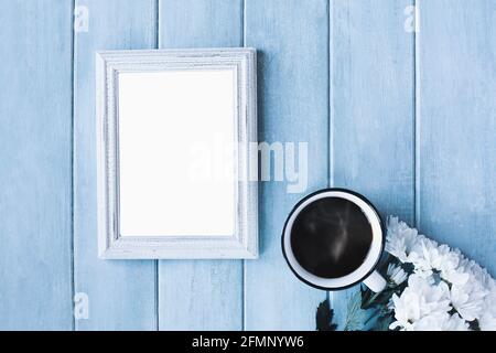 Leerer vertikaler Bilderrahmen auf blauem, rustikalem Hintergrund mit dampfendem Kaffee und weißen Gänseblümchen. Stockfoto