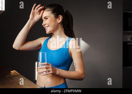 Glückliche junge Brünette Frau in Sportswear hält Glas im Stehen Die Küche Stockfoto