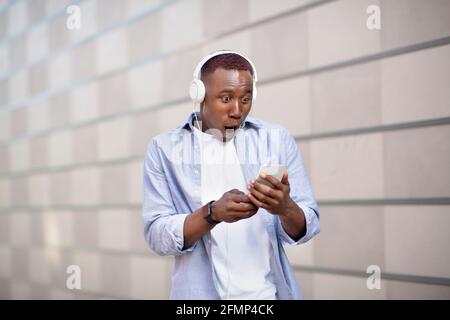 Schockierter afroamerikanischer Mann im Headset starrte auf den Smartphone-Bildschirm An der City Street Stockfoto