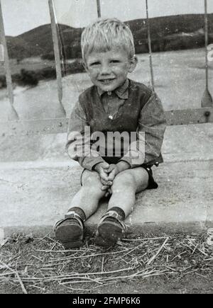 Lächelnder 4-jähriger Junge, der im Dreck auf einer Brücke um 1970, Bulgarien, sitzt Stockfoto