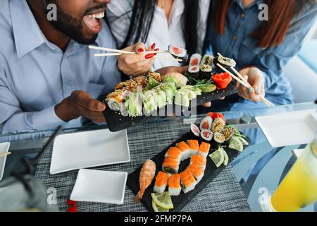 Multirassische Freunde essen Sushi im Café. Leckere Sushi-Brötchen, Maki auf dem Teller serviert. Nahaufnahme von Sushi-Rollen auf dem Teller in den Händen eines afrikanischen Mannes. Zwei Freundinnen, die Rollen in Essstäben halten Stockfoto