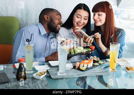 Menschen, Gadgets, Lebensmittel und Blogging-Konzept. Team von multikulturellen jungen Menschen im Café essen Sushi-Brötchen, Spaß haben. Die rothaarige kaukasische Frau macht am Telefon ein Foto von Sushi Stockfoto