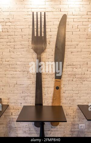 FLORENZ, ITALIEN - 24. Aug 2020: Ein Restauranttisch vor einer sandfarbenen Ziegelwand mit einer übergroßen Gabel und einem Messer, das an der Wand für dec befestigt ist Stockfoto