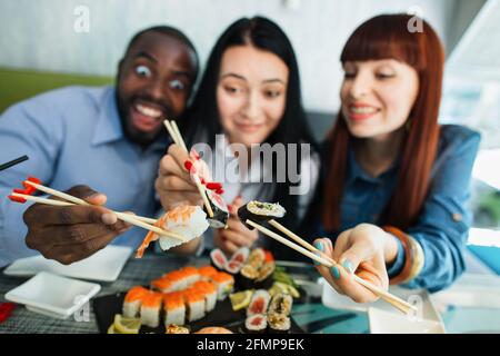 Verschwommener Blick auf lächelnde, angenehme, gemischte Rennfreunde, Spaß beim Mittagessen im chinesischen Restaurant, mit Holzstäben und leckeren Sushi-Brötchen. Konzentrieren Sie sich auf Hände mit Rollen Stockfoto