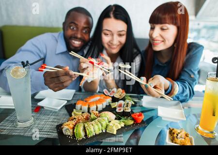 Verschwommener Blick auf lächelnde, angenehme, gemischte Rennfreunde, Spaß beim Mittagessen im chinesischen Restaurant, mit Holzstäben und leckeren Sushi-Brötchen. Konzentrieren Sie sich auf Hände mit Rollen Stockfoto