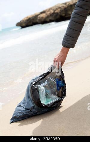 Nahaufnahme eines jungen kaukasischen Mannes, der einen Müllbeutel voller frisch aus dem Sand eines einsamen Strandes gesammelter Abfälle trägt, um die zu säubern Stockfoto