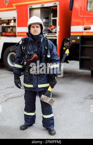 Porträt eines ernsthaften und selbstbewussten kaukasischen Feuerwehrmundes, der im Hintergrund des Staplers eine spezielle Schutzuniform trägt und einen Hammer hält. Stockfoto