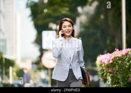 Junge asiatische Geschäftsführerin, die beim Gehen am Handy spricht In der City Street Stockfoto