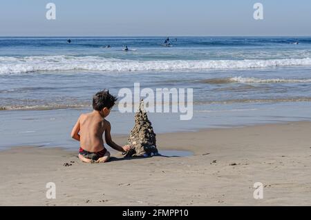 Ein kleiner Junge, der in Venice Beach, Kalifornien, eine Sandburg auf eigene Faust errichtet. Es wurde am Nachmittag genommen. Surfer Dudes sind im Hintergrund zu sehen. Stockfoto