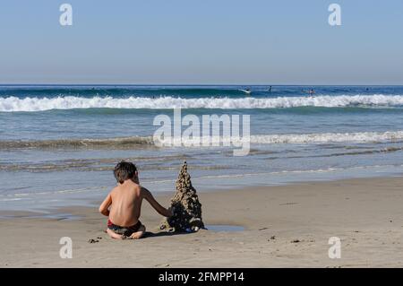 Ein kleiner Junge, der in Venice Beach, Kalifornien, eine Sandburg auf eigene Faust errichtet. Es wurde am Nachmittag genommen. Surfer Dudes sind im Hintergrund zu sehen. Stockfoto