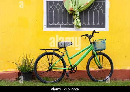 Ein abgenutztes grünes Fahrrad im Freien mit einem niedlichen blauen Korb, der gegen ein gelbes Cottage House gestellt wurde. Stockfoto