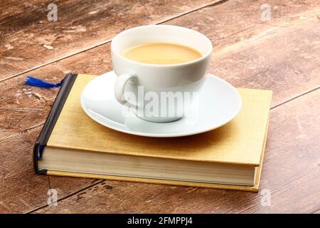 Weiße Tasse und Untertasse mit Tee gefüllt saß auf einem Buch lesen auf einer rustikalen Holztischplatte Stockfoto