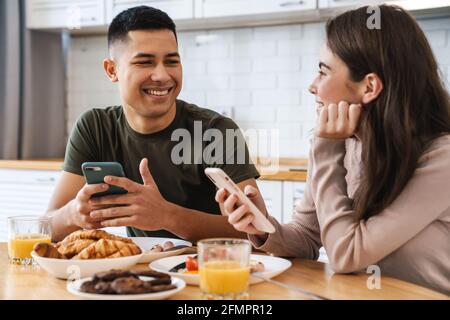 Portrait eines lächelnden Paares mit Smartphones und Gespräch beim Frühstück in der gemütlichen Küche zu Hause Stockfoto