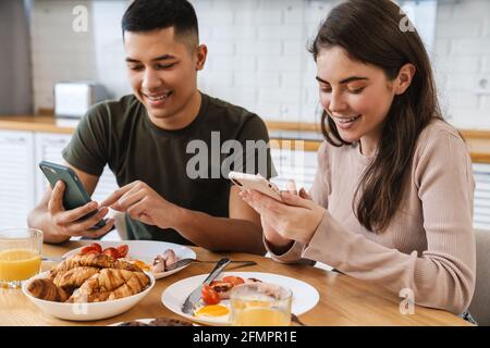 Portrait eines lächelnden Paares mit Smartphones und Gespräch beim Frühstück in der gemütlichen Küche zu Hause Stockfoto
