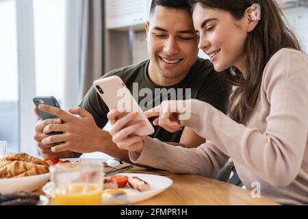 Portrait eines lächelnden Paares mit Smartphones und Gespräch beim Frühstück in der gemütlichen Küche zu Hause Stockfoto