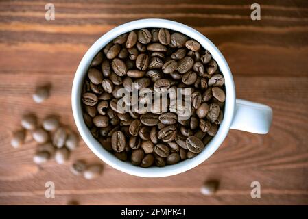 Haufen Kaffeebohnen in weißem Keramikbecher auf Holztisch. Geröstete Kaffeesorten auf Holzoberfläche, Draufsicht Stockfoto