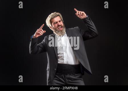 Fröhlicher, junger Geschäftsmann in schwarzem Anzug, der Musik mit Kopfhörern hört und auf grauem Hintergrund tanzt. Stockfoto