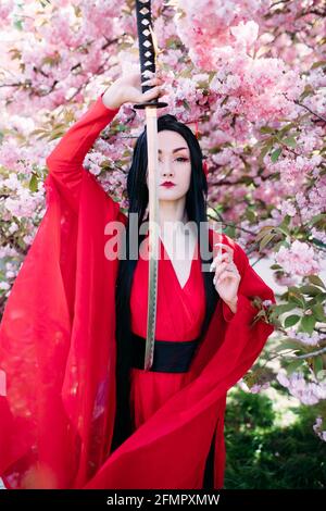 Die junge Frau porträtiert einen traditionellen Kimono in Geisha-Kleidung mit japanischem Samurai-Schwert in der Hand vor dem Hintergrund blühender Sakura-Bäume. Stockfoto