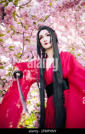 Die junge Frau porträtiert einen traditionellen Kimono in Geisha-Kleidung mit japanischem Samurai-Schwert in der Hand vor dem Hintergrund blühender Sakura-Bäume. Stockfoto