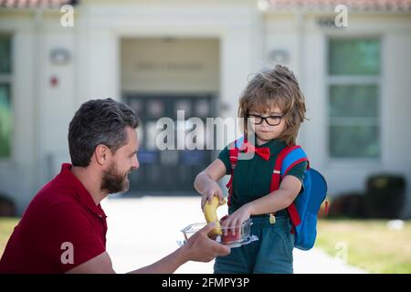 Vater unterstützt und motiviert Sohn. Kind geht zur Grundschule. Kleiner Schuljunge, der im Freien leckeres Mittagessen isst. Lehrertag. Stockfoto